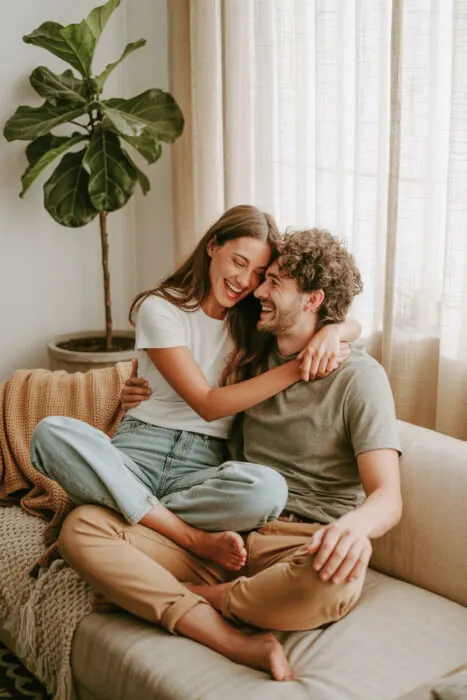 "Couple sitting on a sofa in a cozy living room, laughing together and embracing, wearing casual clothes with warm natural light streaming through curtains – perfect example of casual couple photography at home.