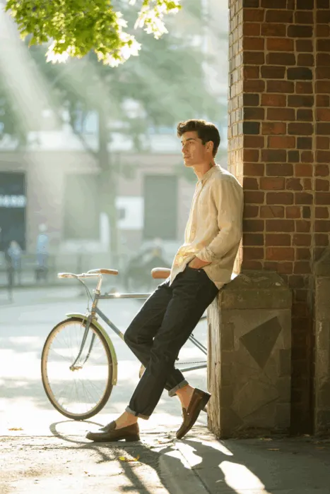 Casual outfit ideas for men: relaxed man in casual shirt and jeans leaning against a brick wall with bicycle nearby, outdoors in sunlight.