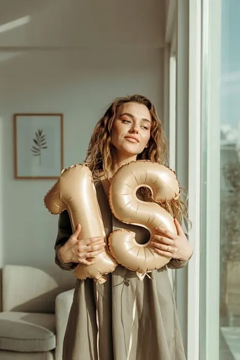 Woman holding large neutral number balloons indoors near a window
