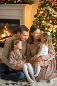Cozy indoor Christmas photo of a family in warm neutral winter clothes sitting near a glowing tree and fireplace in soft golden light