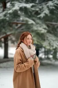 A woman standing outdoors in falling snow wearing a neutral winter coat, photographed in soft natural light with a calm winter mood.