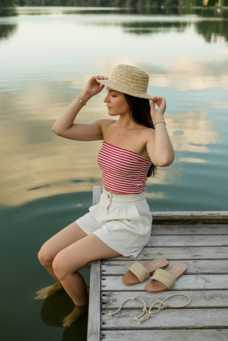 woman in striped bandeau top and white linen shorts sitting on wooden dock over calm lake
